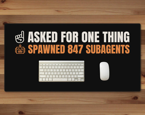 Black 16 x 32 inch desk mat that reads "Asked for one thing / Spawned 847 subagents", on a wood desk with a keyboard and mouse below the joke