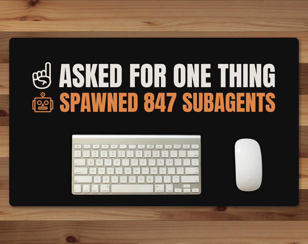 Black 24 x 14 inch desk mat that reads "Asked for one thing / Spawned 847 subagents", on a wood desk with a keyboard and mouse below the joke