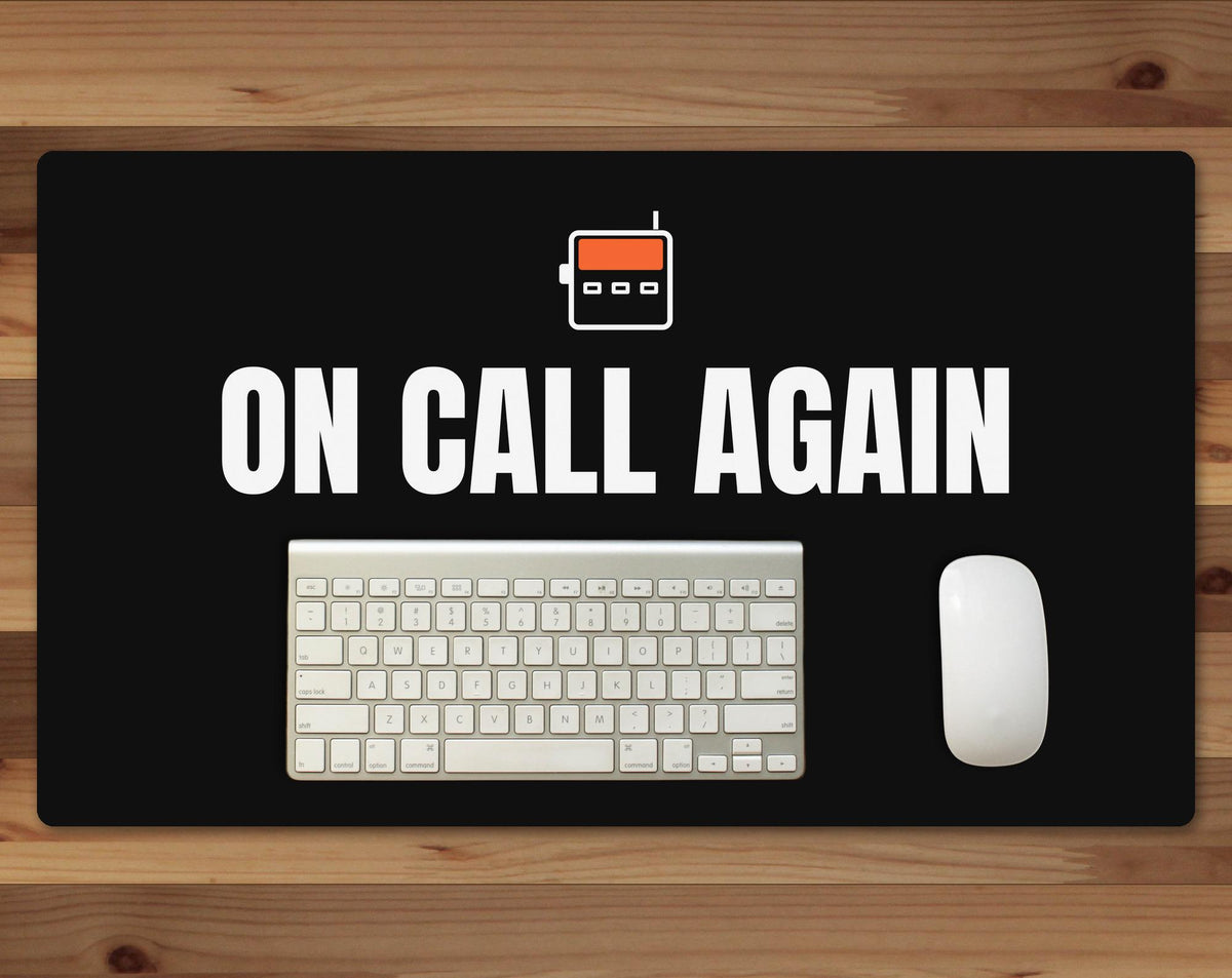 Black 24 x 14 inch desk mat that reads "On call again", on a wood desk with a keyboard and mouse below the joke