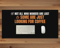 Black 16 x 32 inch desk mat that reads "Not all who wander are lost / Some are just looking for coffee", on a wood desk with a keyboard and mouse below the joke