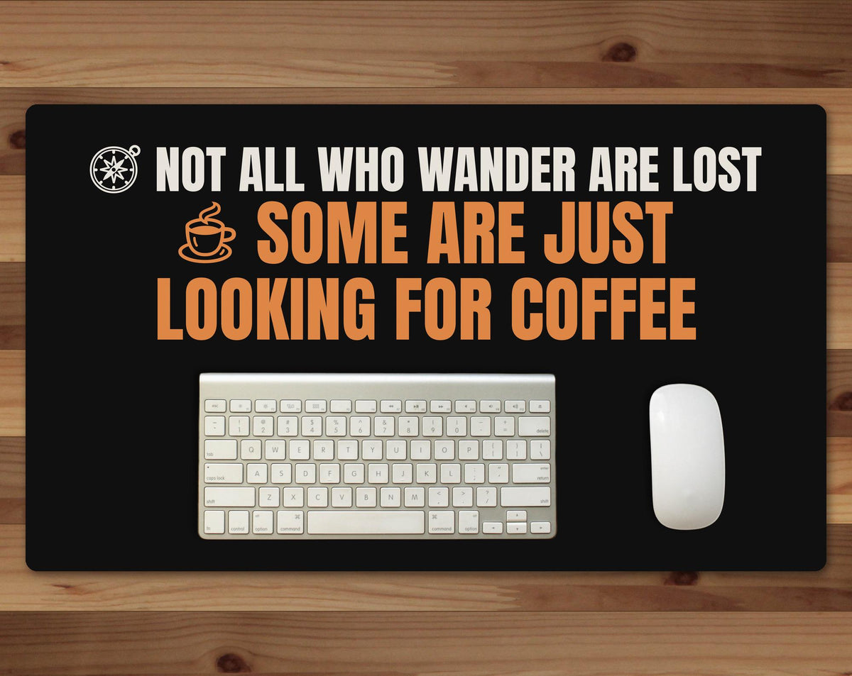 Black 24 x 14 inch desk mat that reads "Not all who wander are lost / Some are just looking for coffee", on a wood desk with a keyboard and mouse below the joke