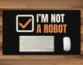 Black 24 x 14 inch desk mat that reads "I’m not / A robot", on a wood desk with a keyboard and mouse below the joke